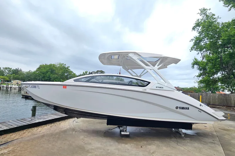 Slide: The Image of 2023 Yamaha Boats 275SDX on dock, sleek design, white exterior, overcast sky. - 2