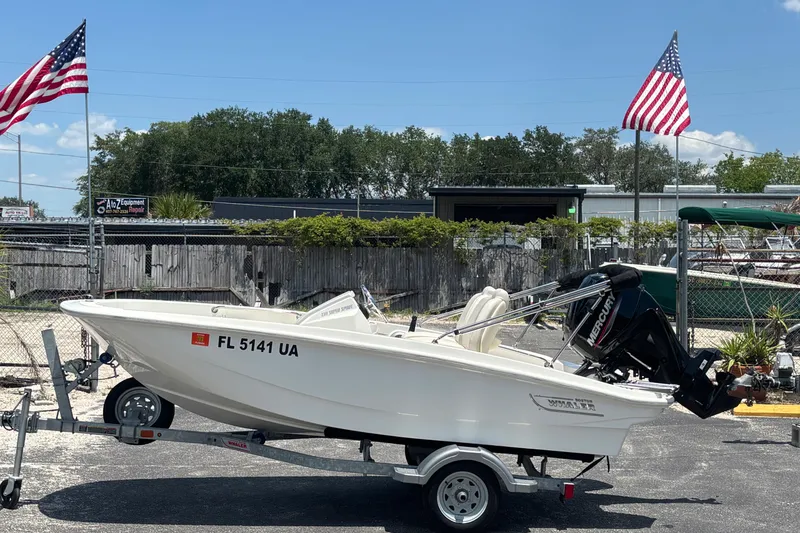 Slide: The Image of 2025 Boston Whaler 130 Super Sport boat on trailer, parked outdoors with American flags. - 7