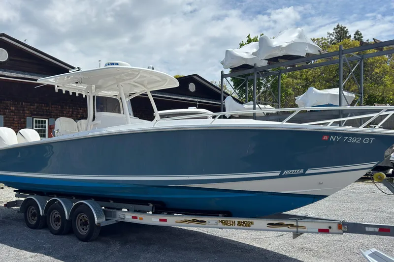 The Image of 2013 Jupiter 32 Cuddy boat on trailer, parked outdoors near wooden building. - 0