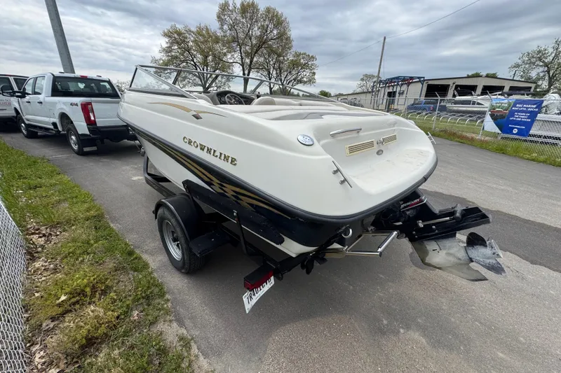 Slide: The Image of 2000 Crownline 180 BR boat on trailer, parked near a white truck, under cloudy skies. - 2