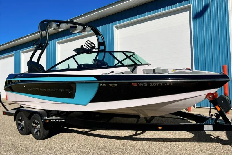 The Image of 2014 Nautique Super Air Nautique 210 boat on trailer, parked near blue building. - 0