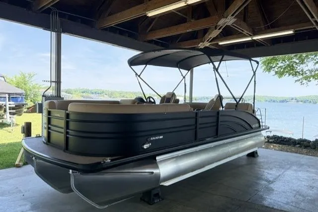 The Image of 2025 Harris Sunliner 230 pontoon boat under a wooden shelter by a scenic lake. - 0