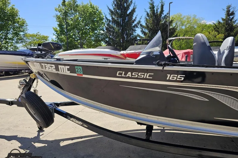 Slide: The Image of 2014 Alumacraft Classic 165 boat on trailer, parked outdoors with trees in background. - 16
