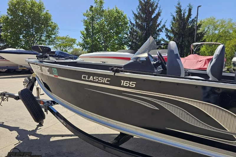 Slide: The Image of 2014 Alumacraft Classic 165 boat on trailer, parked outdoors with trees in background. - 17