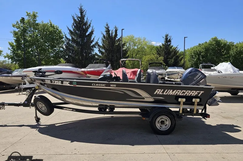 Slide: The Image of 2014 Alumacraft Classic 165 boat on trailer, parked outdoors with other boats. - 18