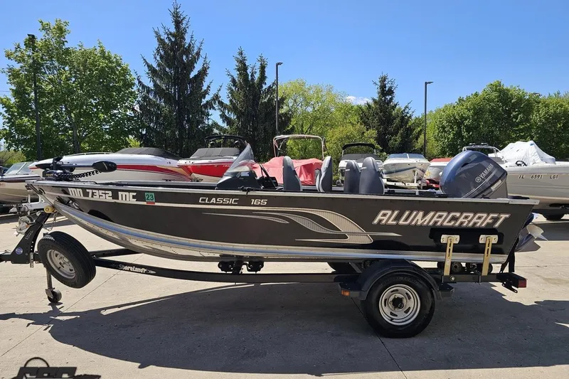 Slide: The Image of 2014 Alumacraft Classic 165 boat on trailer, parked outdoors with trees in background. - 19