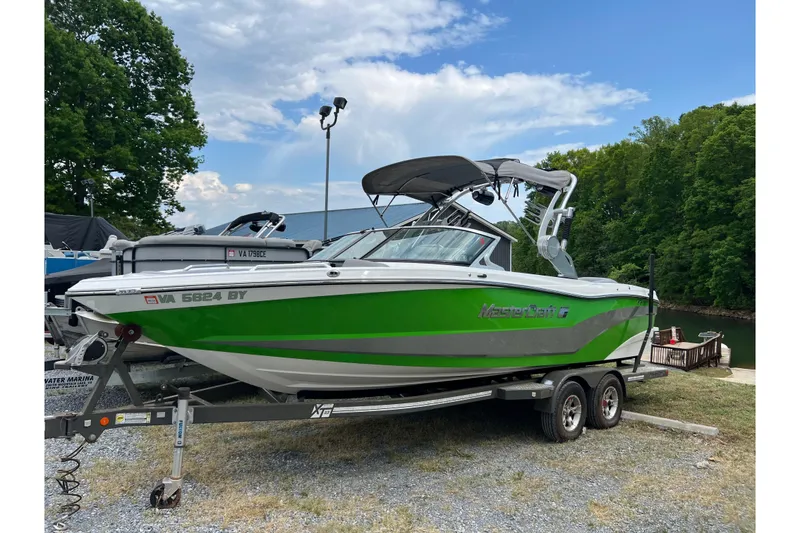 The Image of 2017 MasterCraft XT23 boat in green on a trailer, parked near a lake. - 0