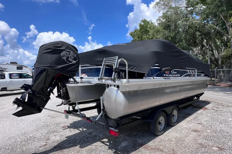Slide: The Image of 2023 Sun Tracker Fishin' Barge 22 DLX pontoon boat on trailer, covered and parked outdoors. - 2
