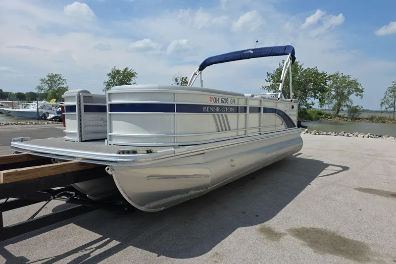 Slide: The Image of 2022 Bennington L 21 L-Bench pontoon boat on trailer by a lakeside. - 2