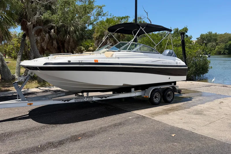 The Image of 2008 NauticStar 210 DC Sport Deck boat on trailer near water, surrounded by trees. - 1