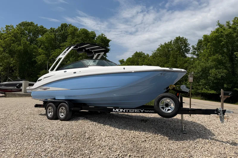 The Image of 2025 Monterey M6 boat on trailer, blue and white design, parked on gravel with trees in background. - 0