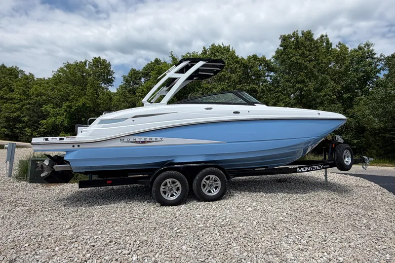 Slide: The Image of 2025 Monterey M6 boat on trailer, blue and white design, parked on gravel with trees in background. - 1