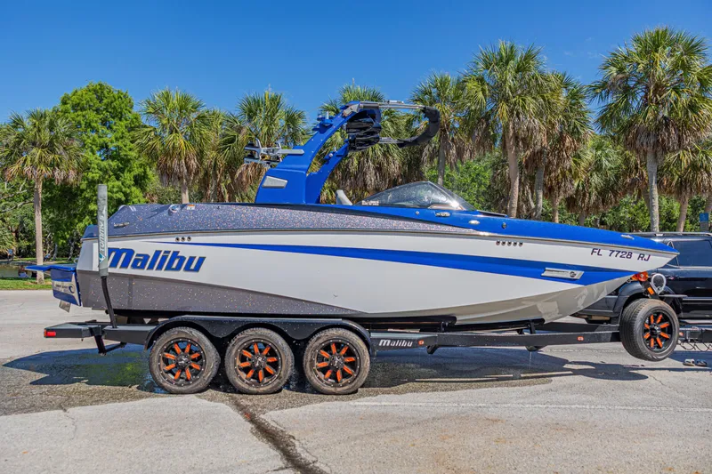 Slide: The Image of 2017 Malibu M235 boat on trailer, blue and white design, parked near palm trees. - 50
