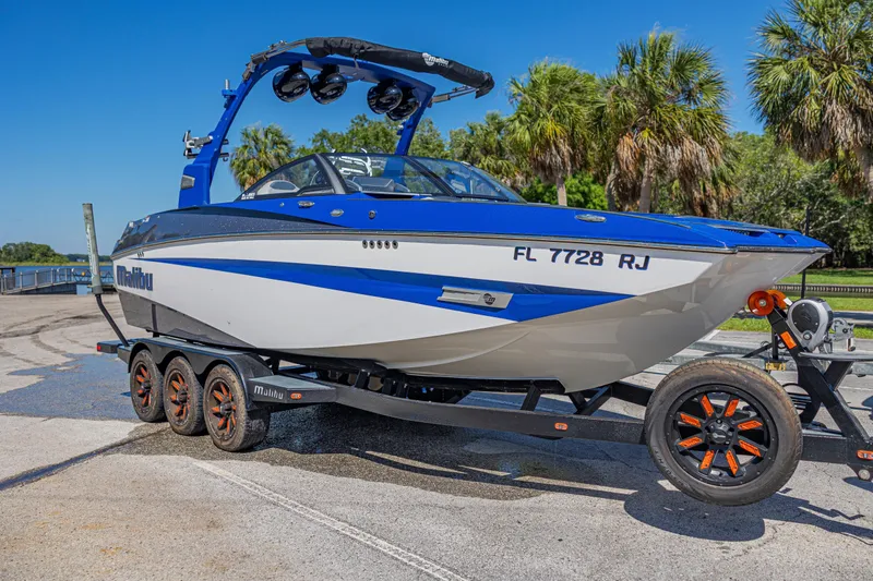 Slide: The Image of 2017 Malibu M235 boat on trailer, blue and white, parked near palm trees. - 51