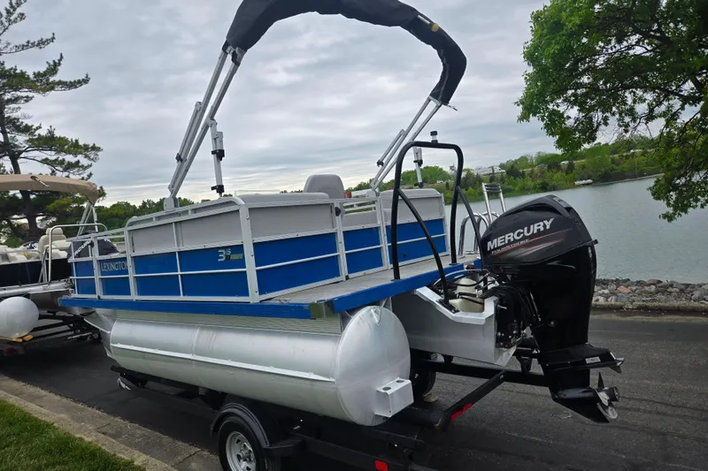 Slide: The Image of 2021 Lexington 315 pontoon boat with Mercury outboard motor on trailer by a lake. - 1