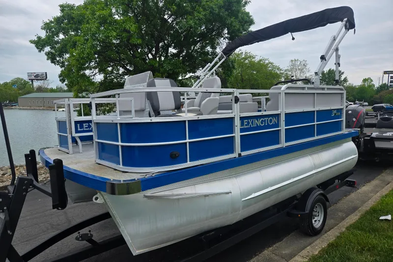 Slide: The Image of 2021 Lexington 315 pontoon boat on trailer by a lakeside, featuring blue and white design. - 9