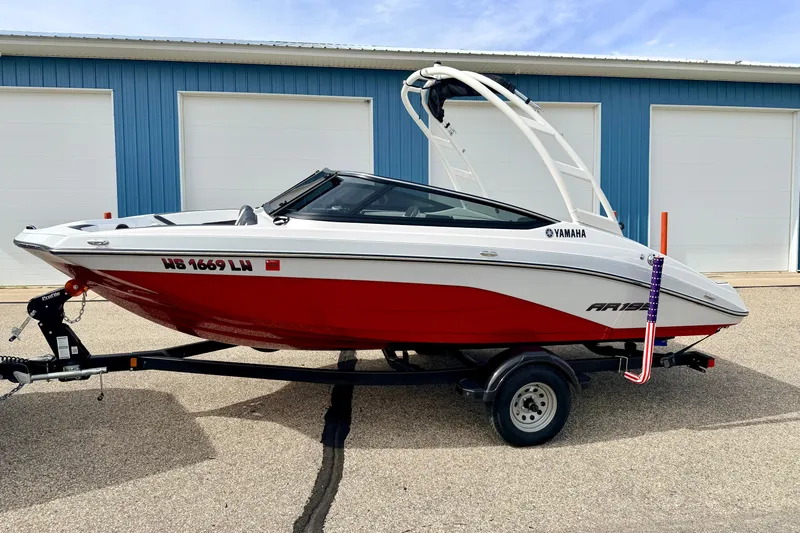 The Image of 2024 Yamaha AR195 boat with red hull on trailer, parked near blue building. - 0