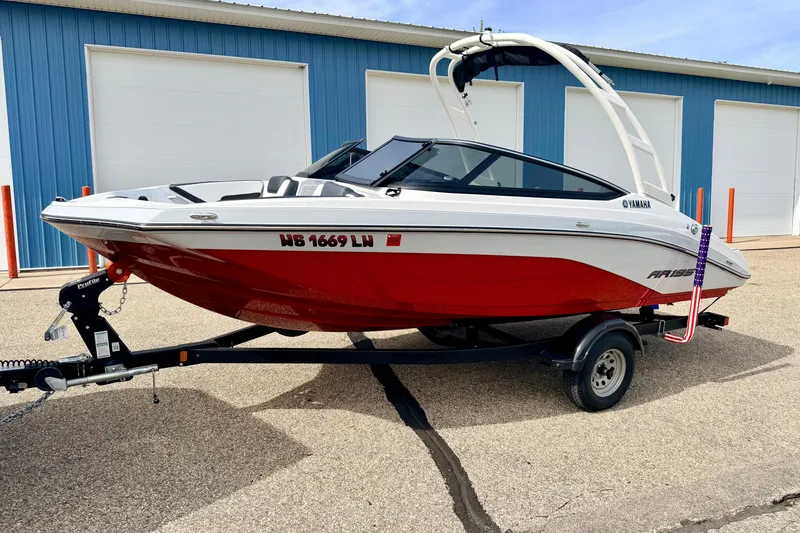Slide: The Image of 2024 Yamaha AR195 boat with red hull on trailer, parked near blue building. - 2