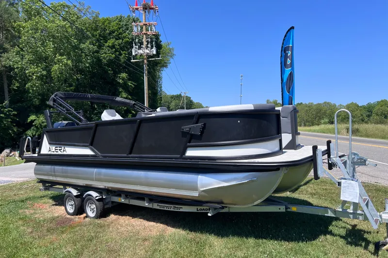 The Image of 2026 Alera M23L pontoon boat on trailer, parked roadside with lush greenery. - 1