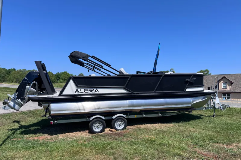 Slide: The Image of Alera M23L 2026 pontoon boat on trailer, parked on grass under clear blue sky. - 2