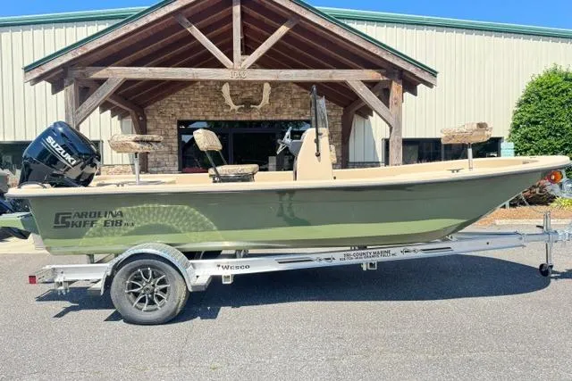 The Image of 2026 Carolina Skiff E18 JVX CC boat on trailer, parked outside a rustic building. - 0