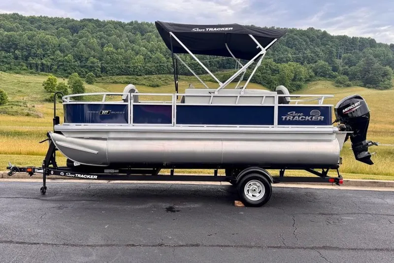 Slide: The Image of 2026 Sun Tracker Bass Buggy 18 DLX pontoon boat on trailer, scenic background. - 1