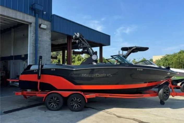 The Image of 2025 MasterCraft NXT23 boat on trailer, black and red design, parked outside a building. - 0