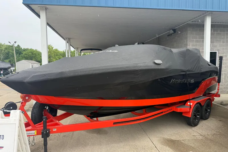 Slide: The Image of 2025 MasterCraft NXT23 boat covered, on a red trailer, parked under a shelter. - 2