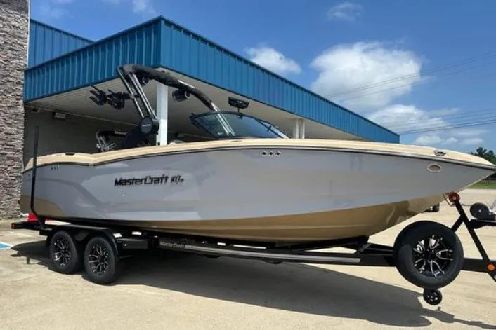 The Image of 2025 MasterCraft NXT24 boat on trailer, parked outside a blue-roofed building under a clear sky. - 0