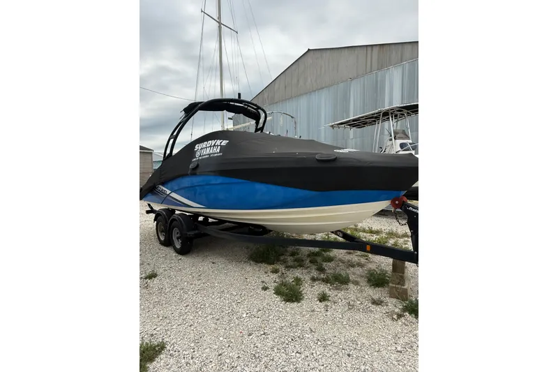 Slide: The Image of 2019 Yamaha Boats AR210 on trailer, covered, parked outdoors near a metal building. - 2