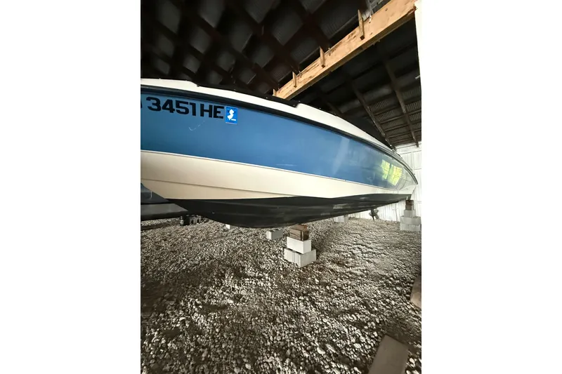 Slide: The Image of 2012 Bayliner 235 Bowrider boat stored indoors on blocks, blue and white hull visible. - 5