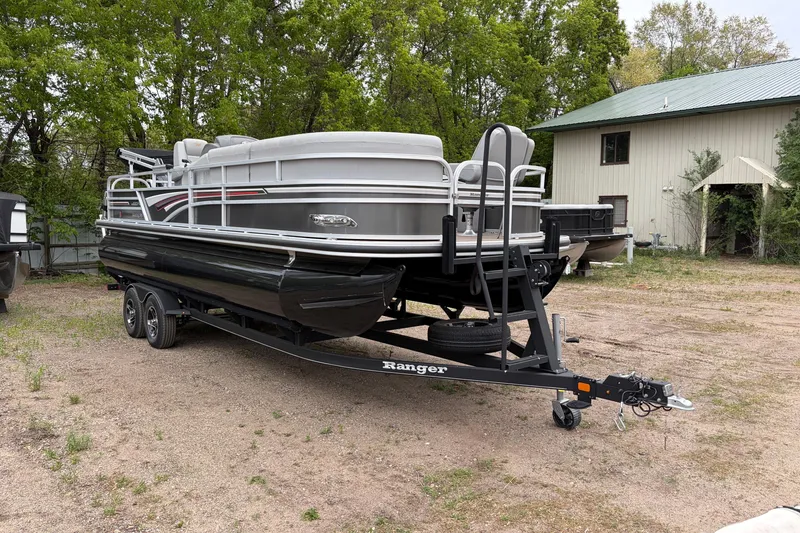 Slide: The Image of 2023 Ranger 220FC pontoon boat on trailer, parked outdoors near trees and building. - 2