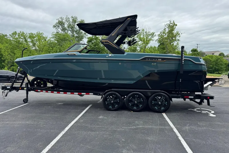 Slide: The Image of 2025 MasterCraft XStar 25 boat on trailer in parking lot, surrounded by trees. - 9