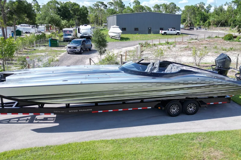 Slide: The Image of 2023 MTI 390X powerboat on trailer, parked outdoors with trees and vehicles in the background. - 2