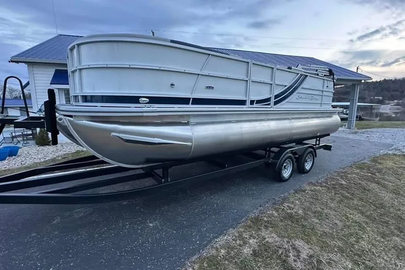Slide: The Image of 2026 South Bay 222CR LE pontoon boat on trailer, parked near a house. - 1
