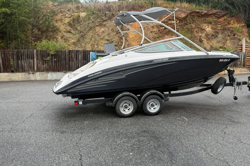 Slide: The Image of 2013 Yamaha AR210 boat on trailer, parked on asphalt, with wooded background. - 2