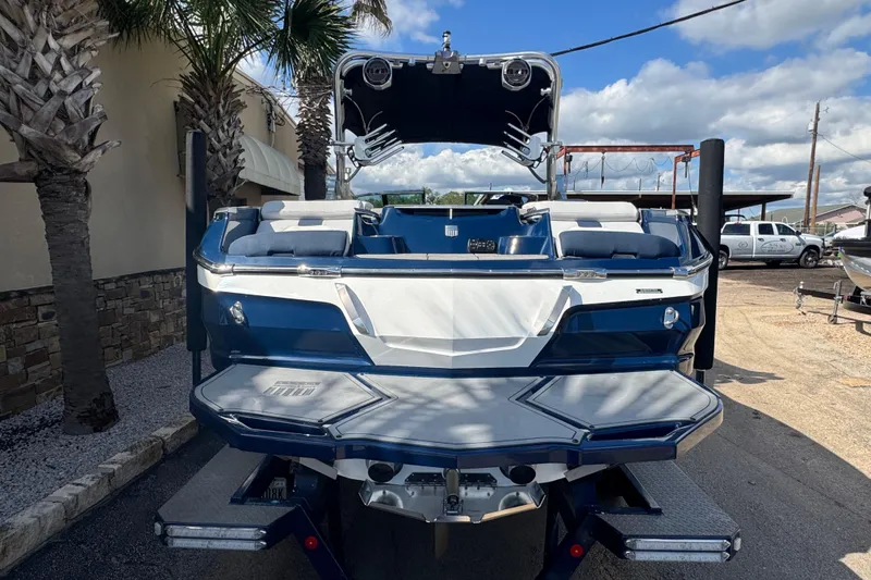 Slide: The Image of 2019 MasterCraft X24 boat, blue and white, parked outdoors with palm trees and vehicles nearby. - 5
