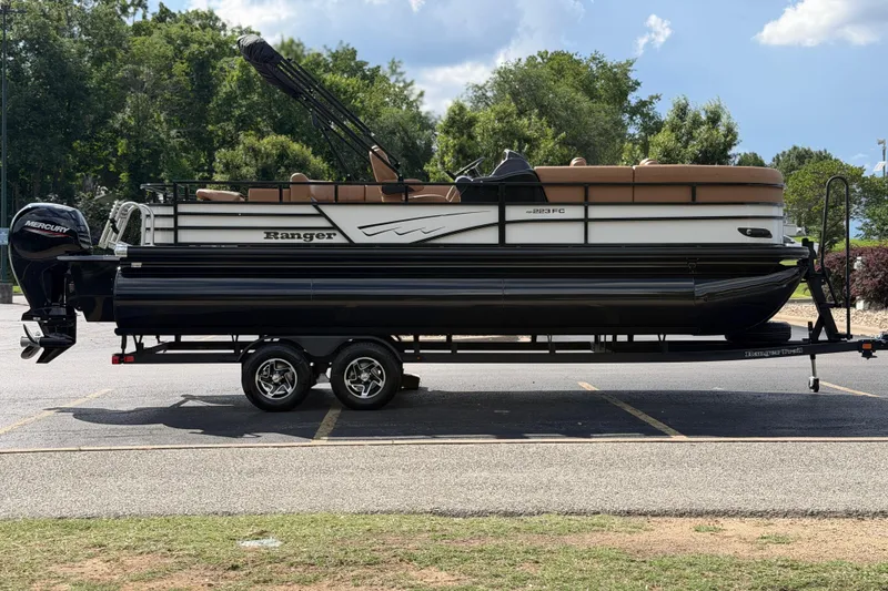 Slide: The Image of 2026 Ranger 223FC pontoon boat on trailer, parked outdoors with trees in background. - 7