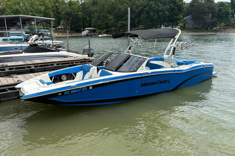 The Image of 2016 MasterCraft X26 boat in blue, docked on a calm lake. - 0