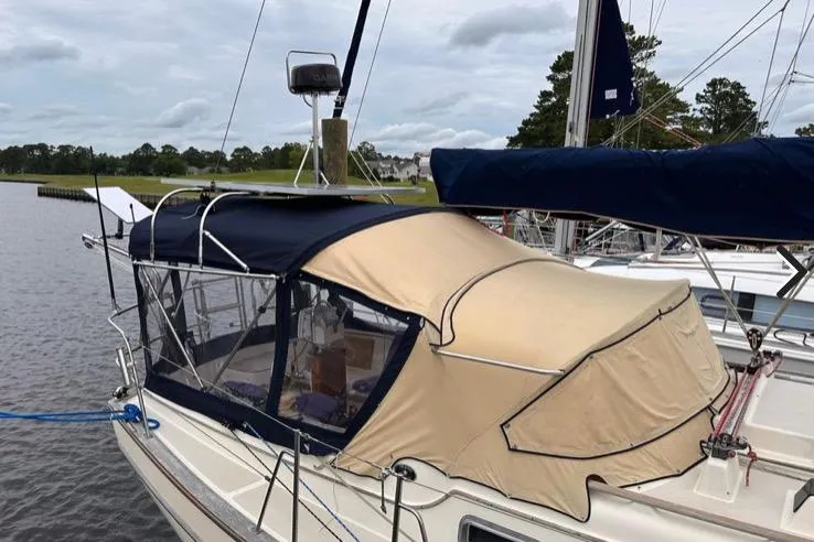 Slide: The Image of 1989 Island Packet 35 sailboat with beige and navy canvas cover docked by a calm river. - 3