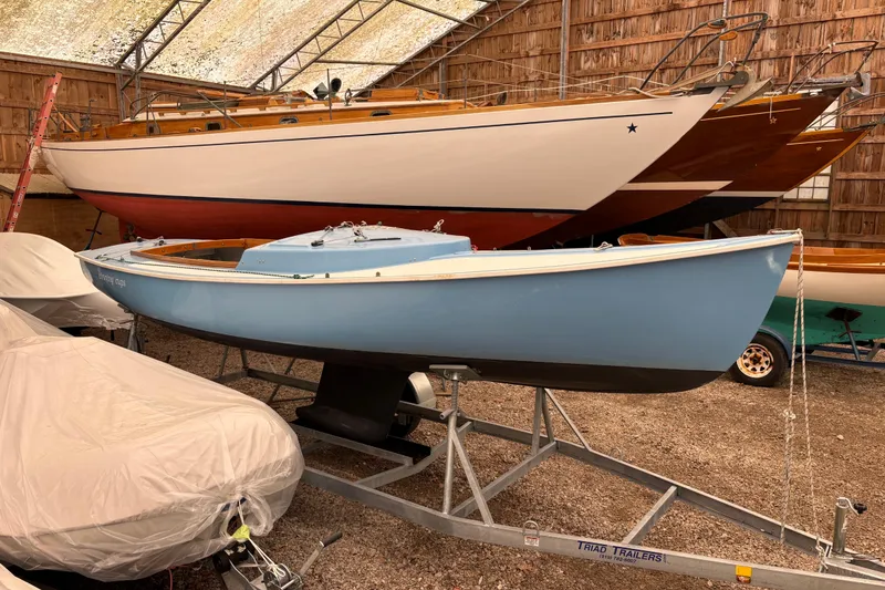 The Image of 1971 Rhodes 19 sailboat in storage, blue hull on trailer, wooden boats in background. - 1