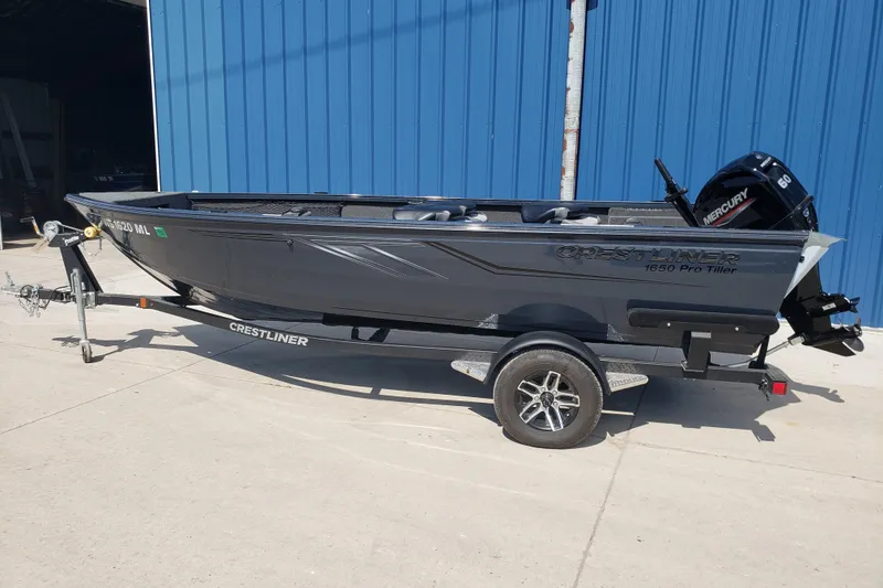 Slide: The Image of 2024 Crestliner 1650 Pro Tiller boat on trailer, parked near blue building. - 2