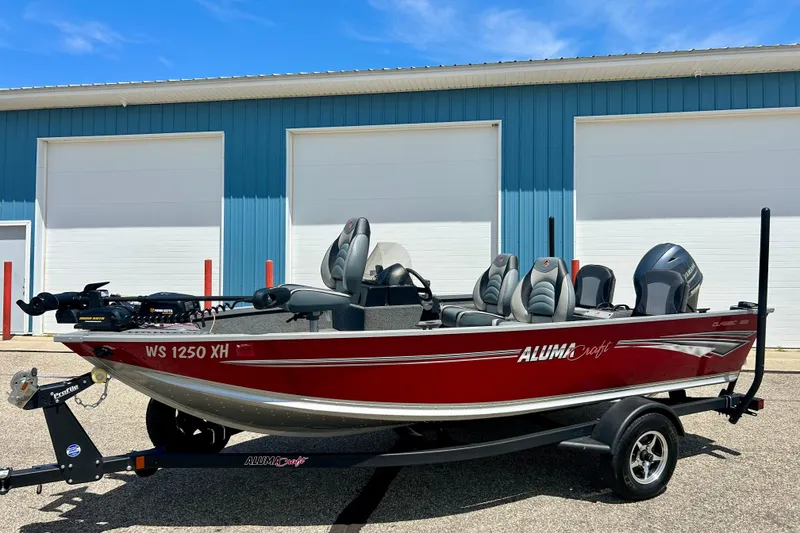 Slide: The Image of 2019 Alumacraft Classic 165 Sport boat in red, parked outside a blue building. - 1