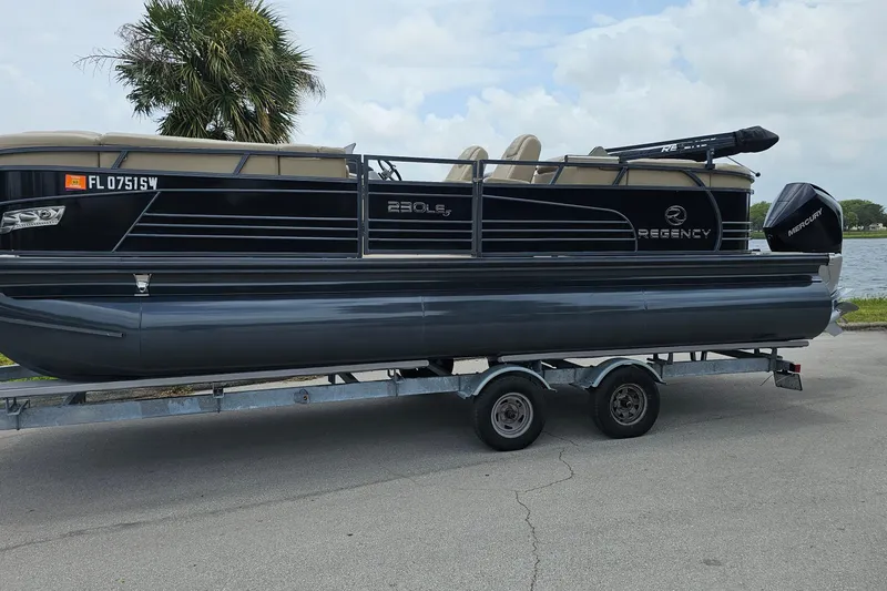 Slide: The Image of 2020 Regency 230 LE3 pontoon boat on trailer, parked near water and palm tree. - 1