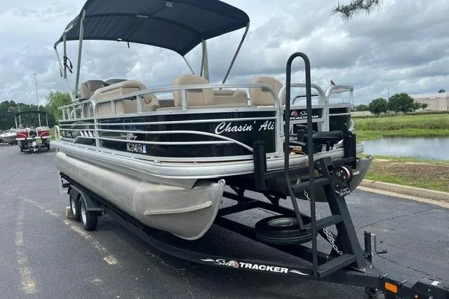 The Image of 2023 SUNTRACKER Fishing Barge 20 pontoon boat on trailer, parked near a lake. - 1
