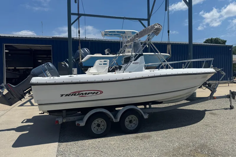 The Image of 2004 Triumph 210 CC boat on trailer, parked outdoors near a blue building. - 0