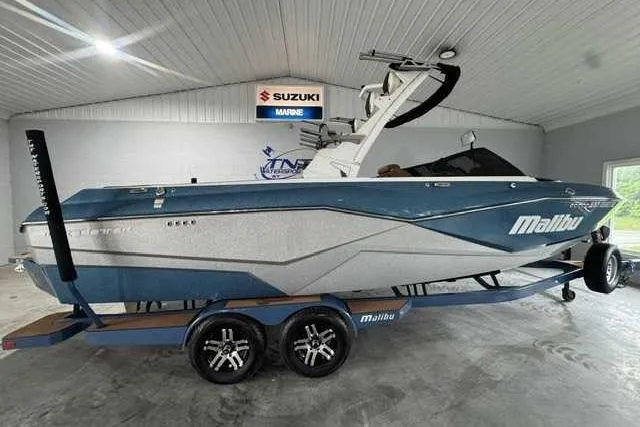 The Image of 2023 Malibu Wakesetter 25 LSV boat in showroom, blue and silver design, on trailer. - 1
