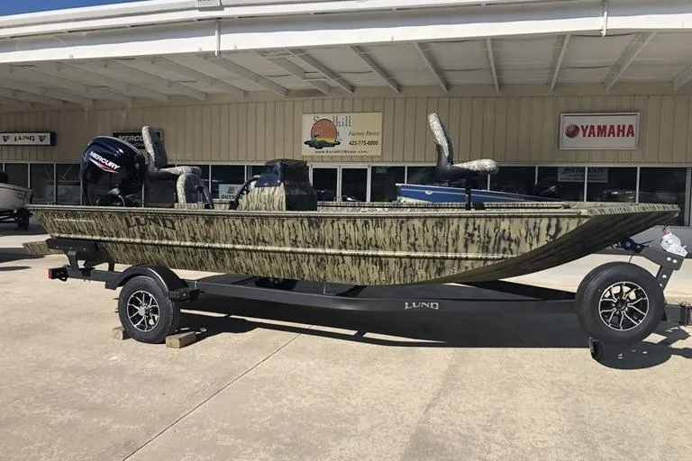 The Image of 2026 Lund 1870 Predator SC boat on trailer, camo design, parked outside dealership. - 1