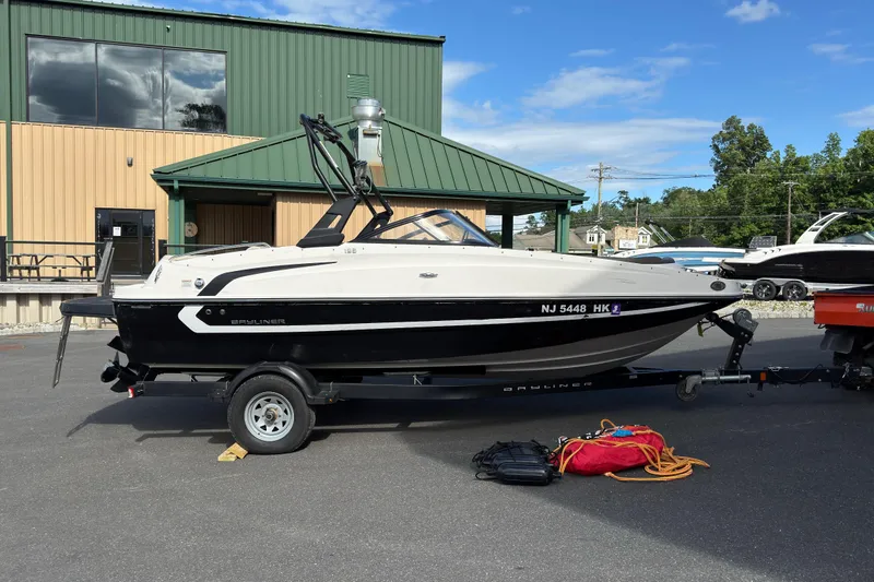 The Image of 2018 Bayliner 195 Deck Boat on trailer, parked outside a green-roofed building. - 0