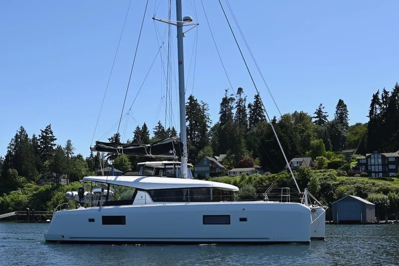 The Image of 2022 Lagoon 42 catamaran sailing near a scenic shoreline with trees and houses. - 0